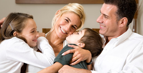 Seated family, dad and daughter looking on whilst the mum hugs son whois kissing her cheek